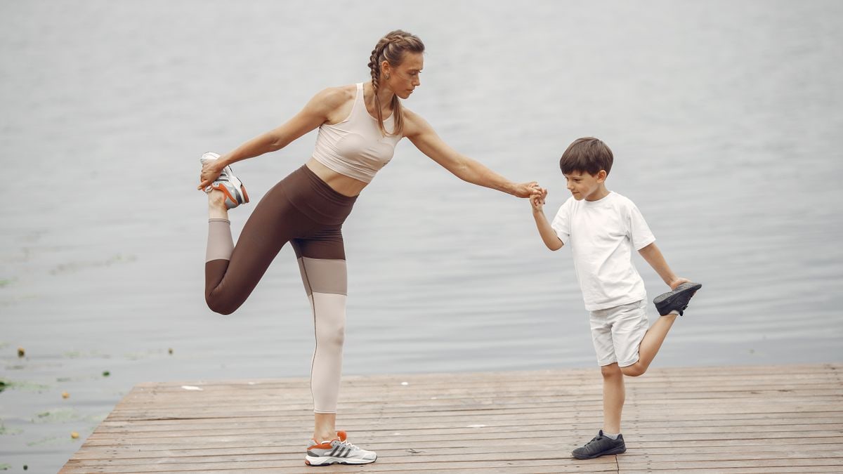 A mother and son holding a yoga pose together on a wooden pier by the water, promoting family fitness.